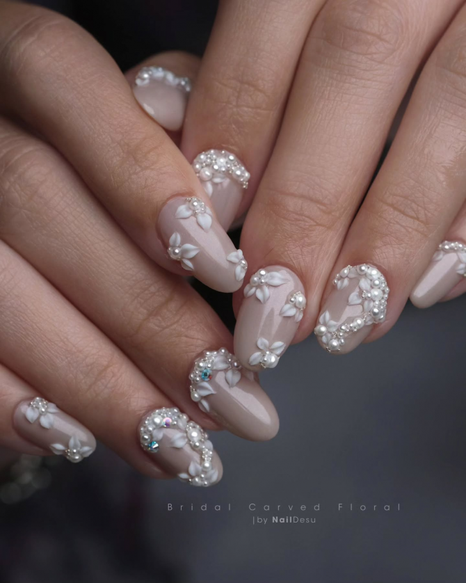 Almond-shaped bridal nails with nude polish, 3D carved white flowers, pearls, and rhinestone accents in an elegant floral design.