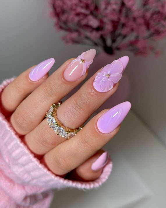 Almond-shaped nails in pastel pink with glossy finish, featuring 3D lavender flower accents for a romantic spring manicure.