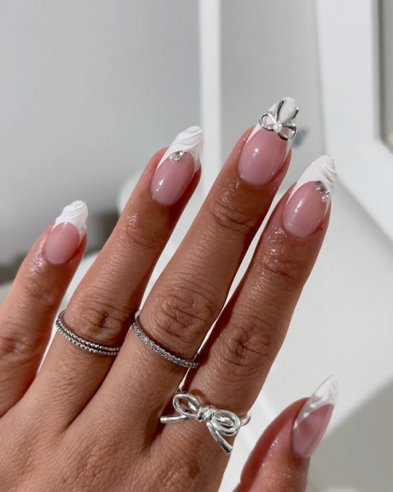 Almond nails with nude pink base, white marble French tips, and 3D silver bow accent
