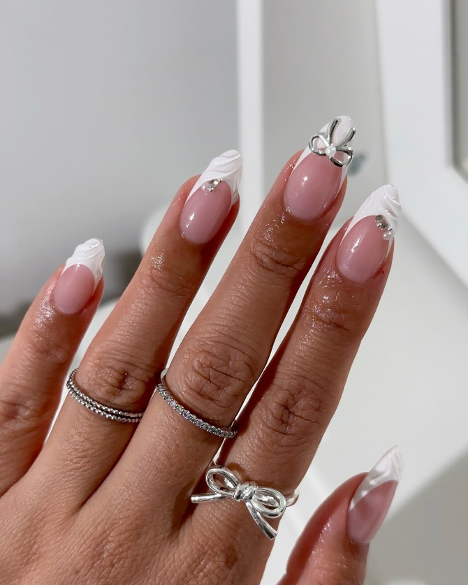 Almond nails with nude pink base, white marble French tips, and 3D silver bow accent