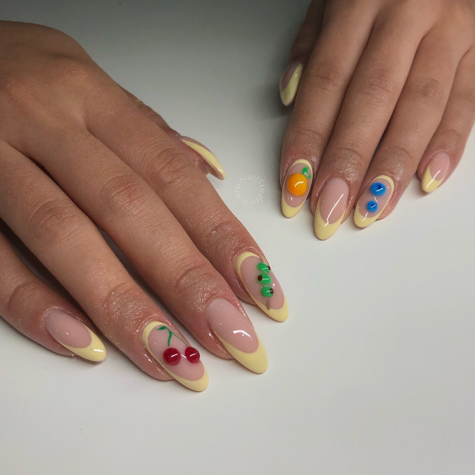 Almond nails with nude base, yellow French tips, and 3D sculpted fruits like cherries, oranges, and blueberries