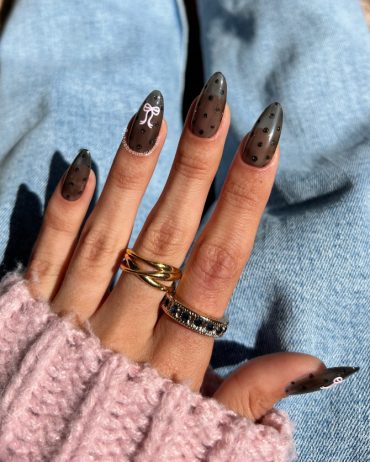 Almond-shaped nails with sheer black polish, tiny black glitter dots, and a white bow design for a Parisian-inspired manicure
