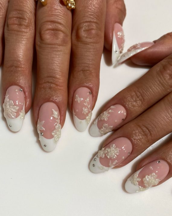 Almond-shaped bridal nails with nude pink base, white French tips, 3D floral accents, and rhinestones