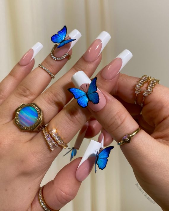 Long square nails with nude base, white French tips, and 3D electric blue butterfly decorations