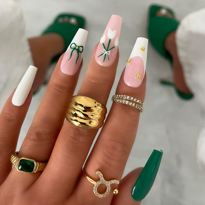 Long coffin nails with white and nude pink base, tulip and bow art, and gold stud embellishments 