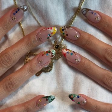 Almond-shaped nude nails with glossy colorful French tips and multicolored gemstone accents for a playful, artistic look