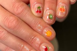 Glossy short round nails with clear base and tiny fruit art designs including cherry, orange, kiwi, blueberry, lemon, and watermelon