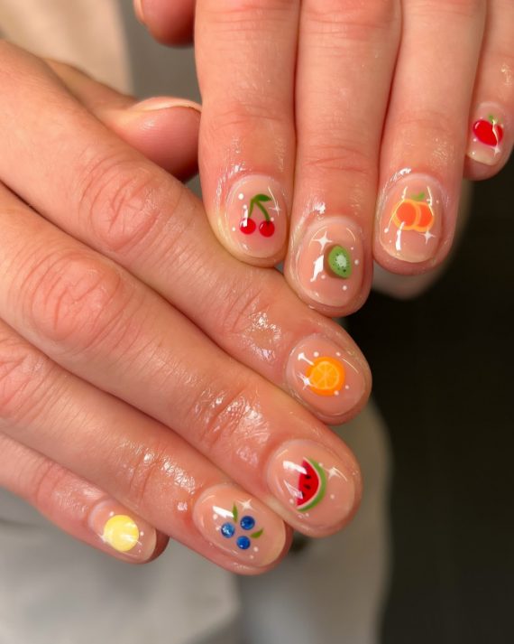 Glossy short round nails with clear base and tiny fruit art designs including cherry, orange, kiwi, blueberry, lemon, and watermelon