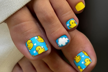Toe nails with blue background featuring yellow rubber ducks, white clouds, tiny hearts, and matte yellow accents on smaller toes.