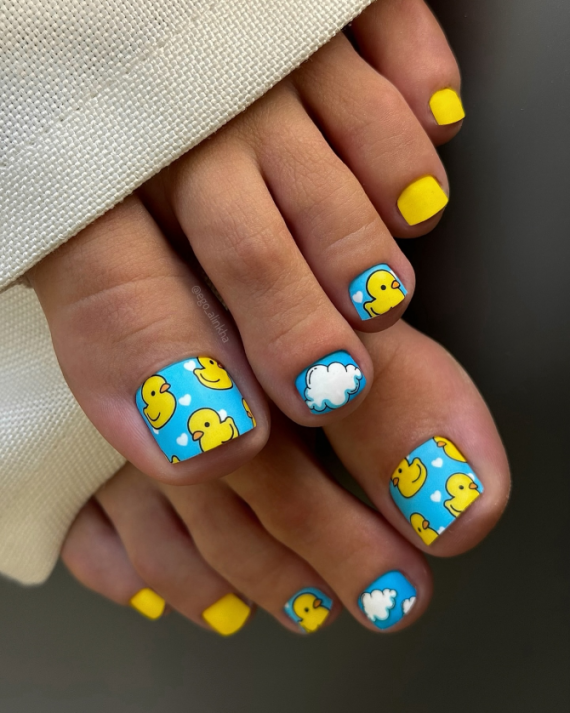 Toe nails with blue background featuring yellow rubber ducks, white clouds, tiny hearts, and matte yellow accents on smaller toes.