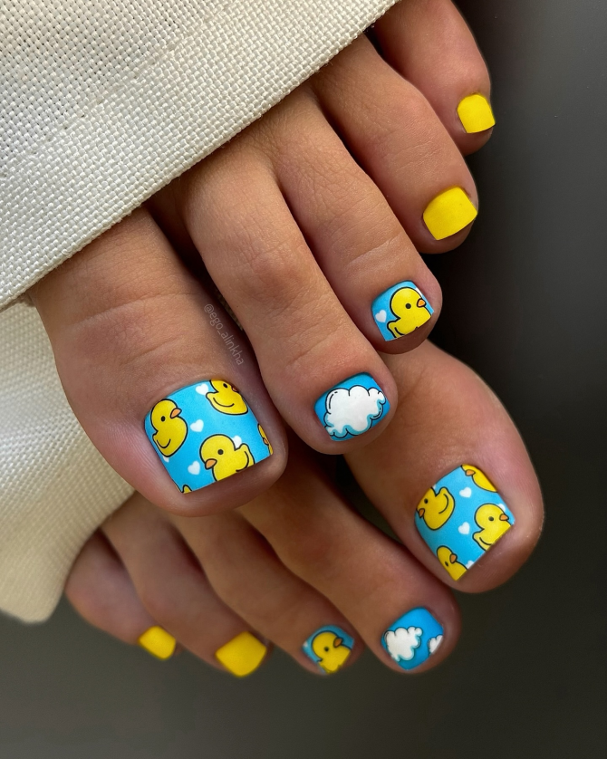 Toe nails with blue background featuring yellow rubber ducks, white clouds, tiny hearts, and matte yellow accents on smaller toes.