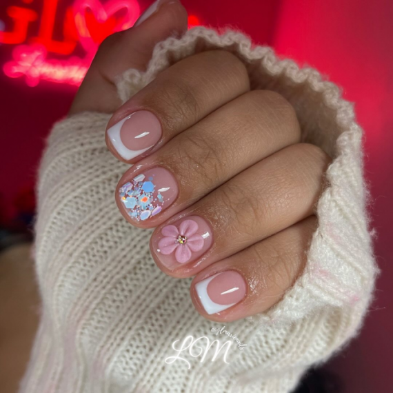 Short square nails with classic white French tips, one with pink 3D flower accent and another with colorful glitter flakes on a nude base.