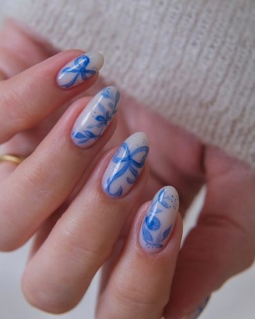 Oval-shaped nails with a nude base and intricate blue floral and bow designs, inspired by porcelain art
