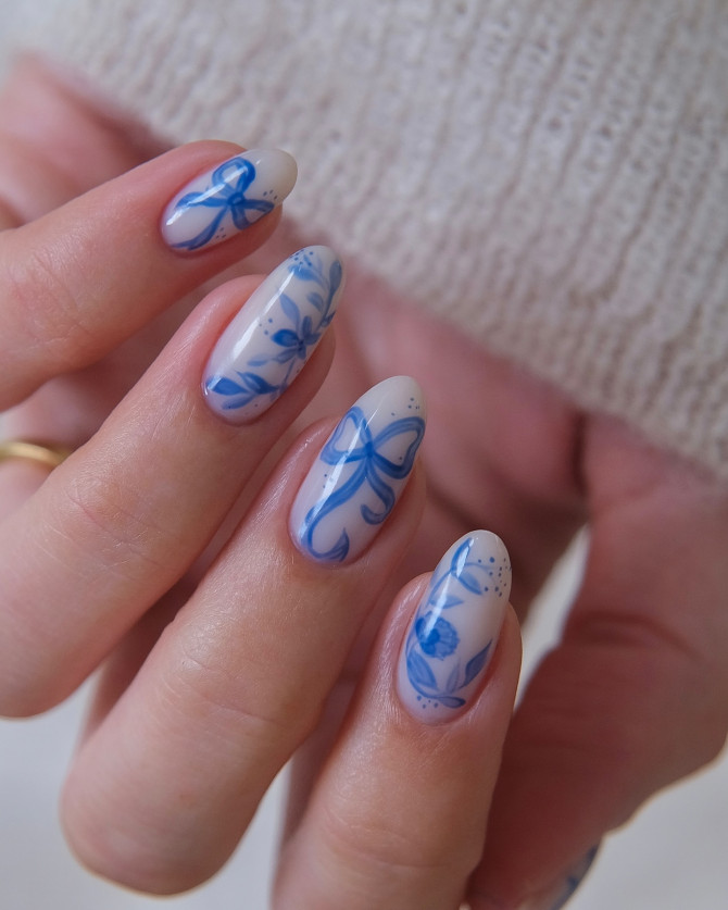 Oval-shaped nails with a nude base and intricate blue floral and bow designs, inspired by porcelain art