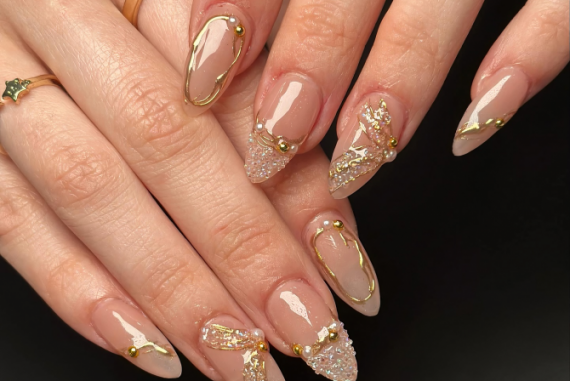 Almond-shaped nude nails with gold chrome outline, gold beads, and crystal glitter accents on a glossy finish.