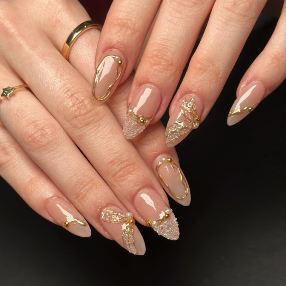 Almond-shaped nude nails with gold chrome outline, gold beads, and crystal glitter accents on a glossy finish.