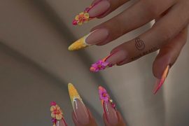 Stiletto-shaped nails with nude base, neon orange and yellow tips, and 3D flower decorations in pink and yellow.