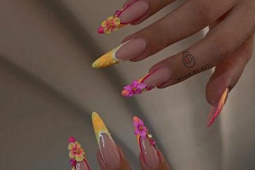 Stiletto-shaped nails with nude base, neon orange and yellow tips, and 3D flower decorations in pink and yellow.