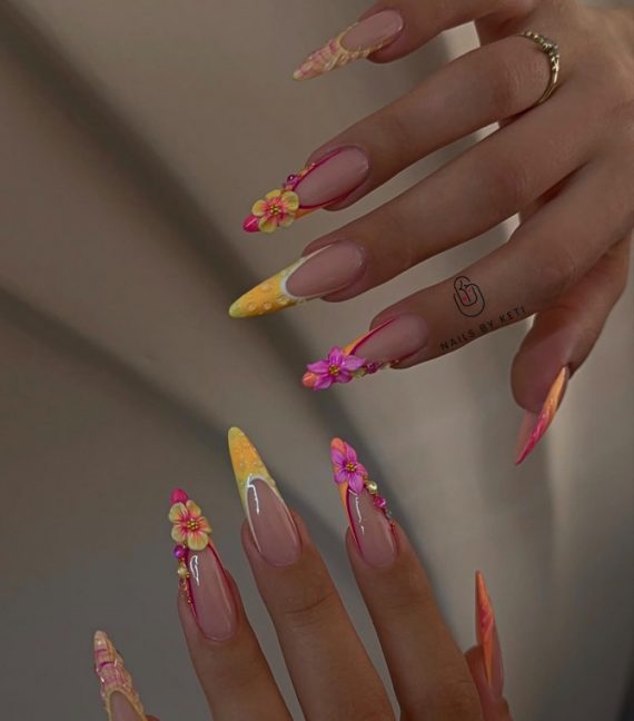 Stiletto-shaped nails with nude base, neon orange and yellow tips, and 3D flower decorations in pink and yellow.