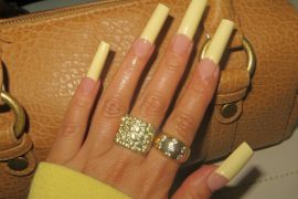 Extra long coffin-shaped nails with glossy yellow French tips and a nude base, styled with bold gold rings