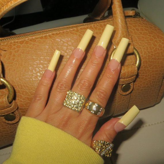 Extra long coffin-shaped nails with glossy yellow French tips and a nude base, styled with bold gold rings