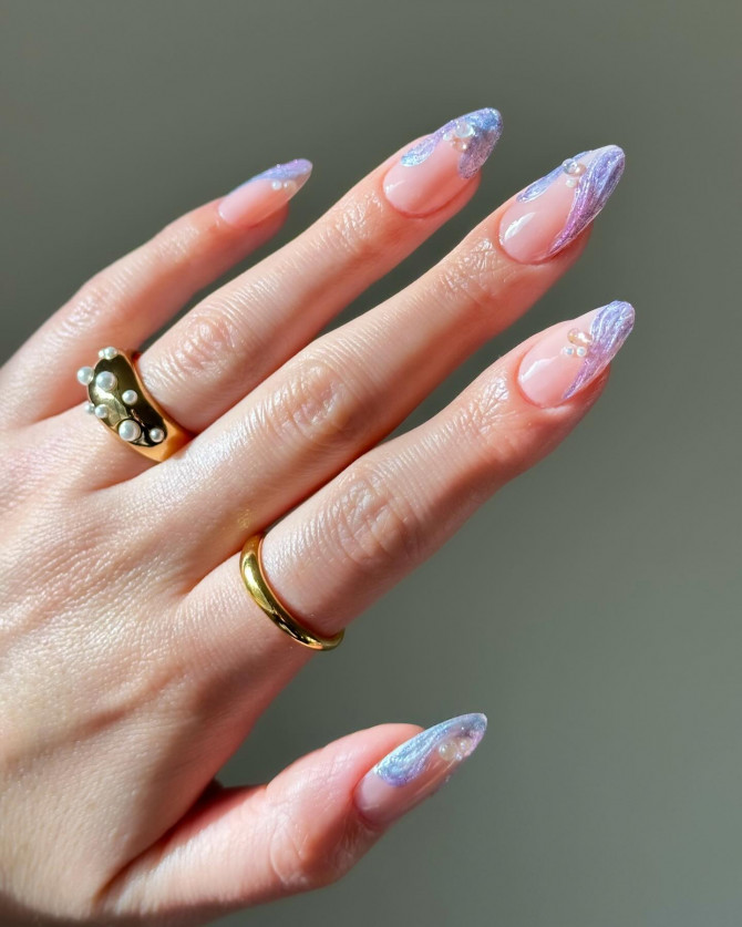 Almond-shaped nails with nude base, iridescent lilac waves, and small pearl accents