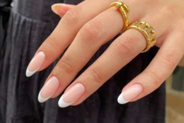 Almond-shaped nails with nude pink base and crisp white French tips, styled with gold rings