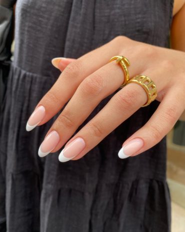 Almond-shaped nails with nude pink base and crisp white French tips, styled with gold rings