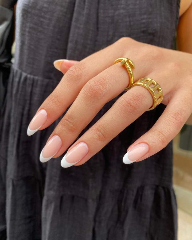 Almond-shaped nails with nude pink base and crisp white French tips, styled with gold rings