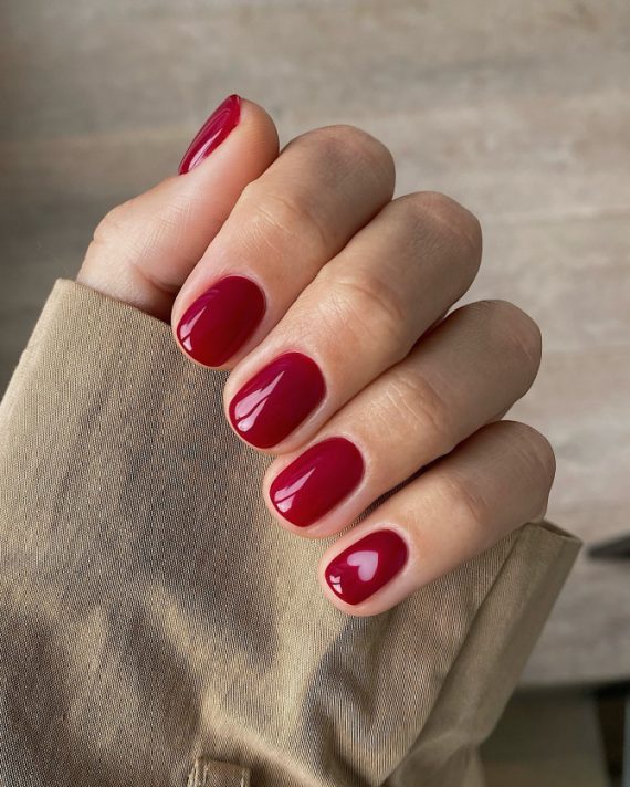 Short round nails painted in glossy cherry red with a small heart-shaped negative space detail on the ring finger