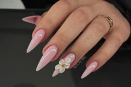 Long stiletto nails in glossy nude pink with a 3D white flower accent on the ring finger, featuring gold center detail.