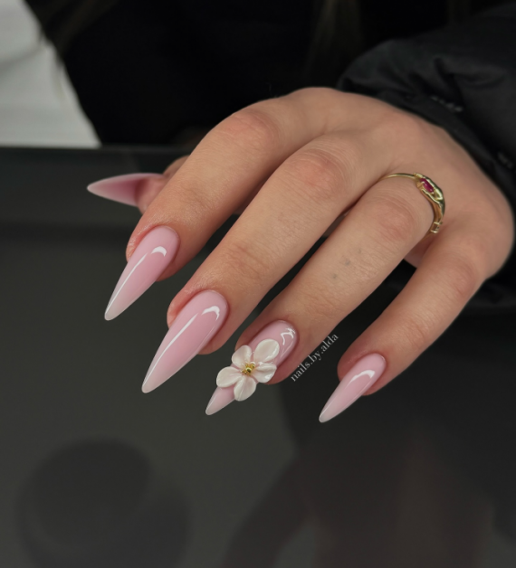 Long stiletto nails in glossy nude pink with a 3D white flower accent on the ring finger, featuring gold center detail.