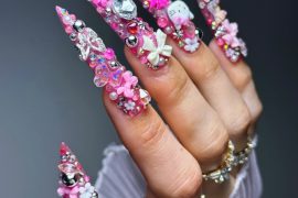 Hand with long pink stiletto nails featuring Hello Kitty charms, crystals, bows, and 3D embellishments in an extravagant kawaii nail design.