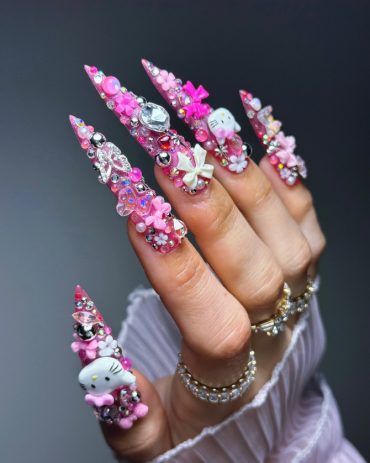 Hand with long pink stiletto nails featuring Hello Kitty charms, crystals, bows, and 3D embellishments in an extravagant kawaii nail design.