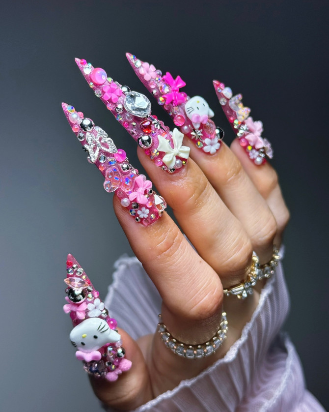 Hand with long pink stiletto nails featuring Hello Kitty charms, crystals, bows, and 3D embellishments in an extravagant kawaii nail design.