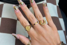 Almond-shaped nude nails with 3D metallic gold drip design and multicolored gem accents