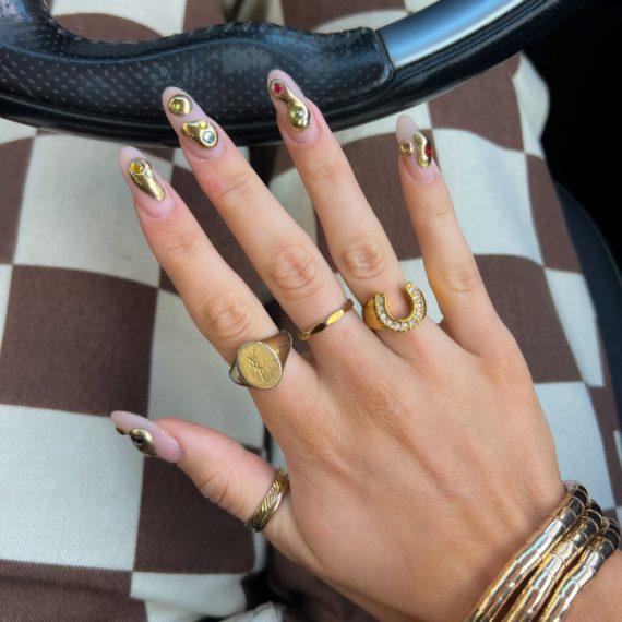 Almond-shaped nude nails with 3D metallic gold drip design and multicolored gem accents