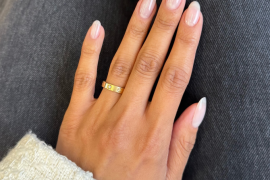 Natural almond-shaped nails with a clear glossy top coat, worn on a hand with a gold ring and white knit sleeve.