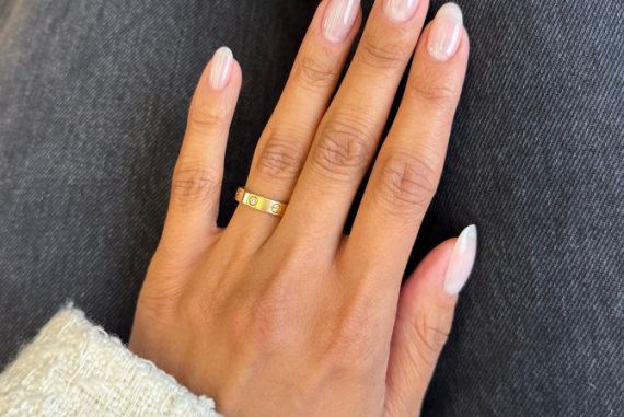 Natural almond-shaped nails with a clear glossy top coat, worn on a hand with a gold ring and white knit sleeve.