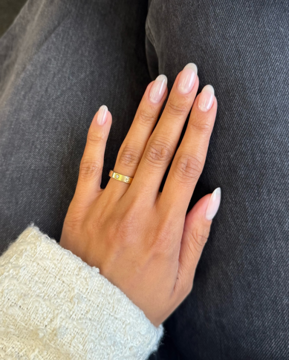 Natural almond-shaped nails with a clear glossy top coat, worn on a hand with a gold ring and white knit sleeve.