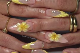 Long stiletto nails with nude base, sculpted yellow textured tips, 3D yellow flowers, and rhinestone accents