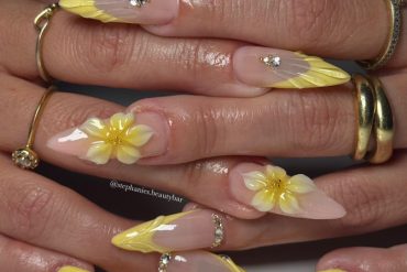Long stiletto nails with nude base, sculpted yellow textured tips, 3D yellow flowers, and rhinestone accents