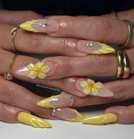 Long stiletto nails with nude base, sculpted yellow textured tips, 3D yellow flowers, and rhinestone accents
