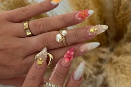 Almond-shaped nails with ombre tips, 3D pink flowers, seashell details, and gold accents, styled for a tropical beach-inspired look.