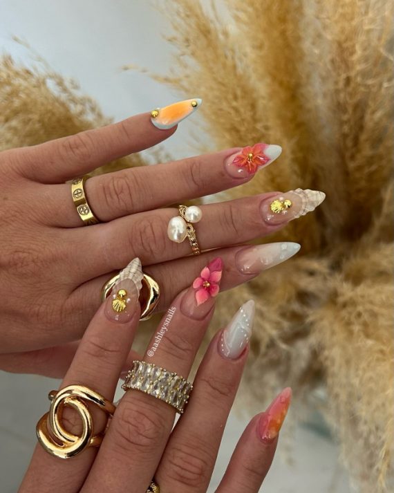 Almond-shaped nails with ombre tips, 3D pink flowers, seashell details, and gold accents, styled for a tropical beach-inspired look.