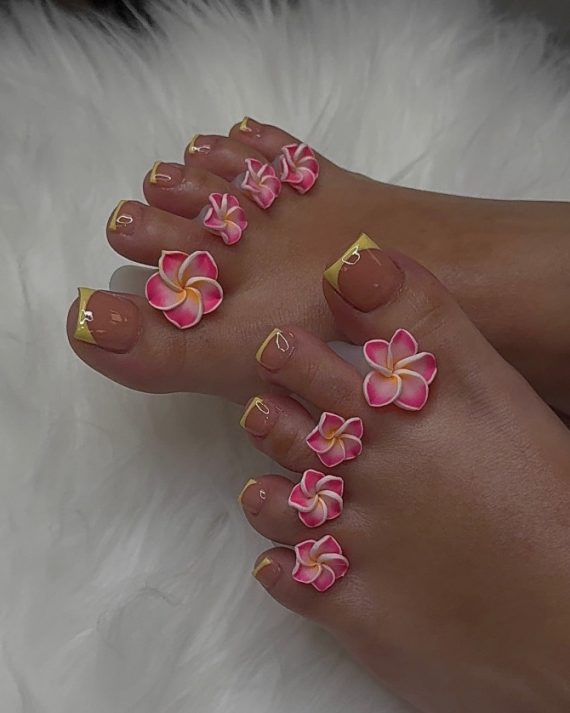 Toenails with nude polish and yellow French tips, decorated with pink and white 3D plumeria flowers on each toe.