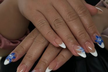 Almond nails with nude base, white French tips, blue floral designs, and soft wave accents