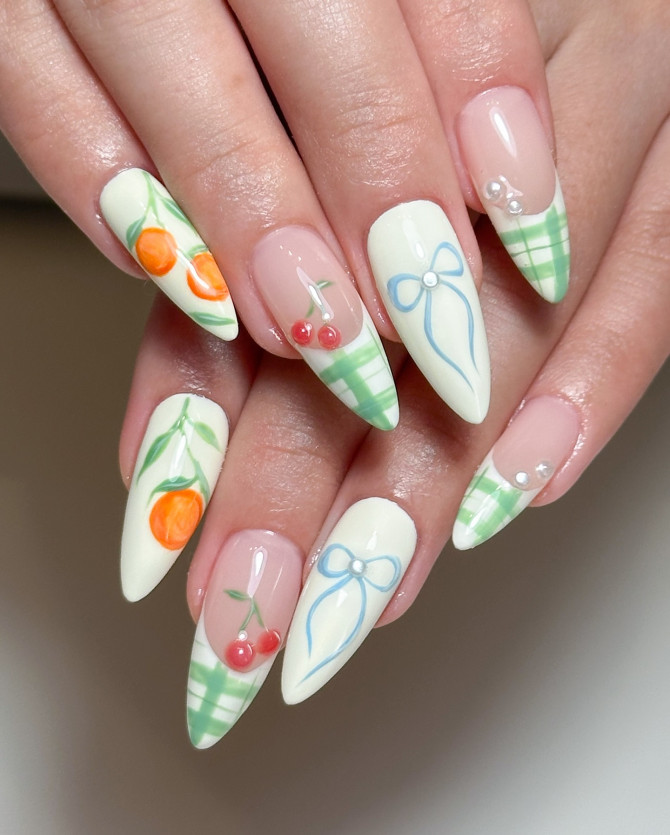 Almond-shaped pastel nails with cherries, bows, gingham tips, and orange fruit accents on a creamy white base