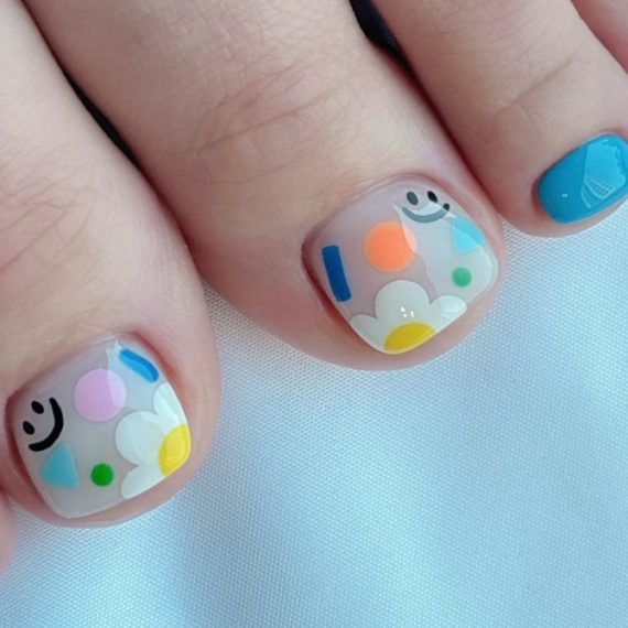 Toe nails with translucent base featuring smiley faces, colorful abstract shapes, and daisy flowers for a cheerful design