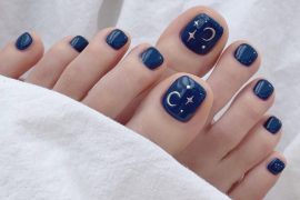 Square-shaped navy blue pedicure with crescent moon and star art on the big toes, perfect for a celestial summer nail look.